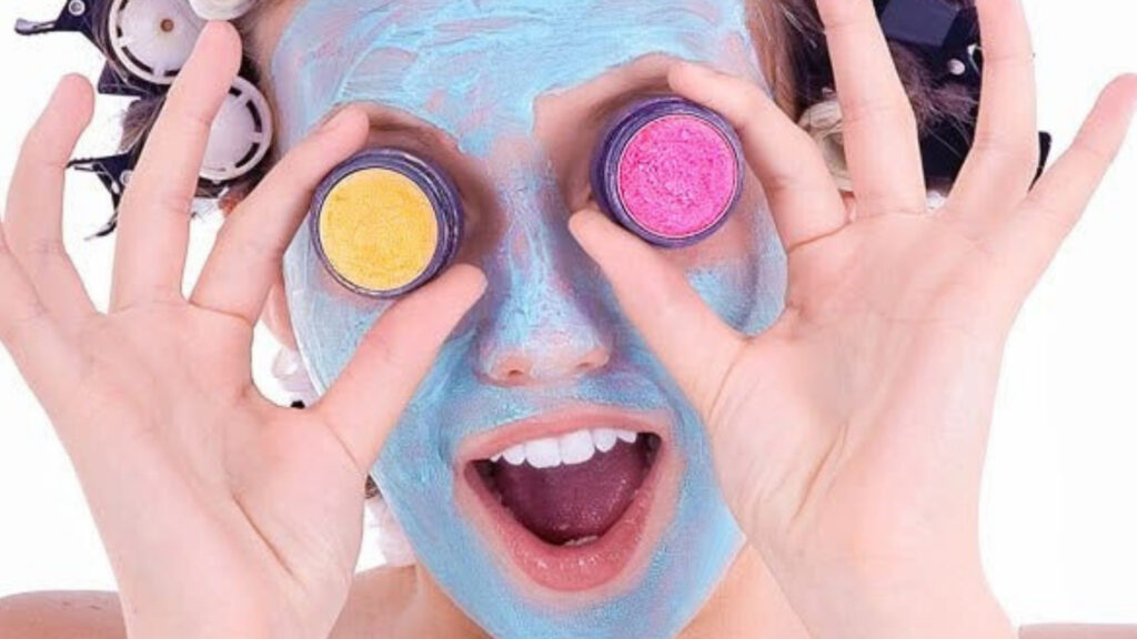 model with blue face mask on her face and eye shadow pots held by her eyes.